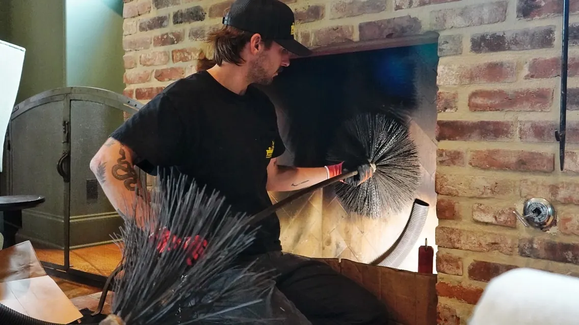 A technician holding a chimney sweeping brush beside a brick fireplace