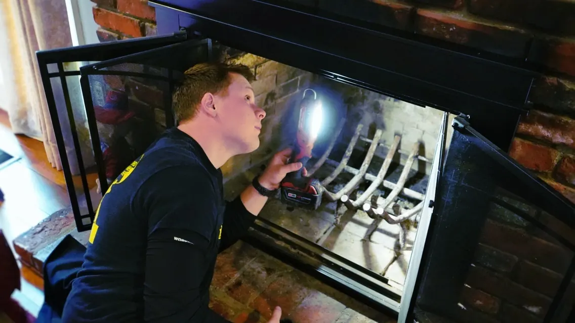 A technician inspecting the firebox of a glass-front fireplace with a work light