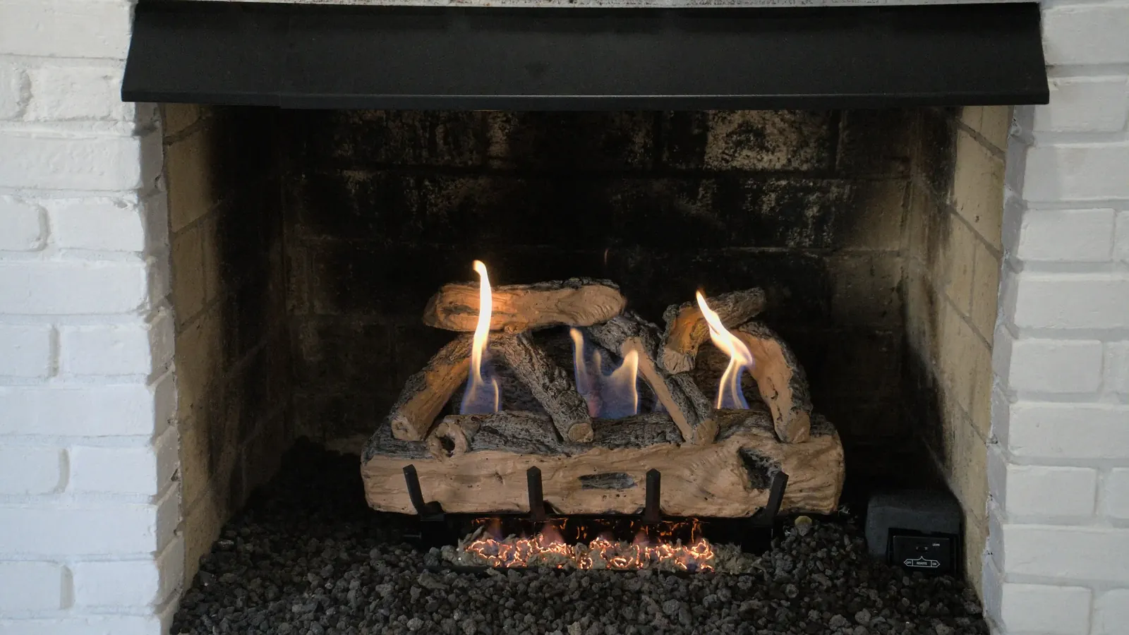 A wide white brick fireplace with a lit gas log set