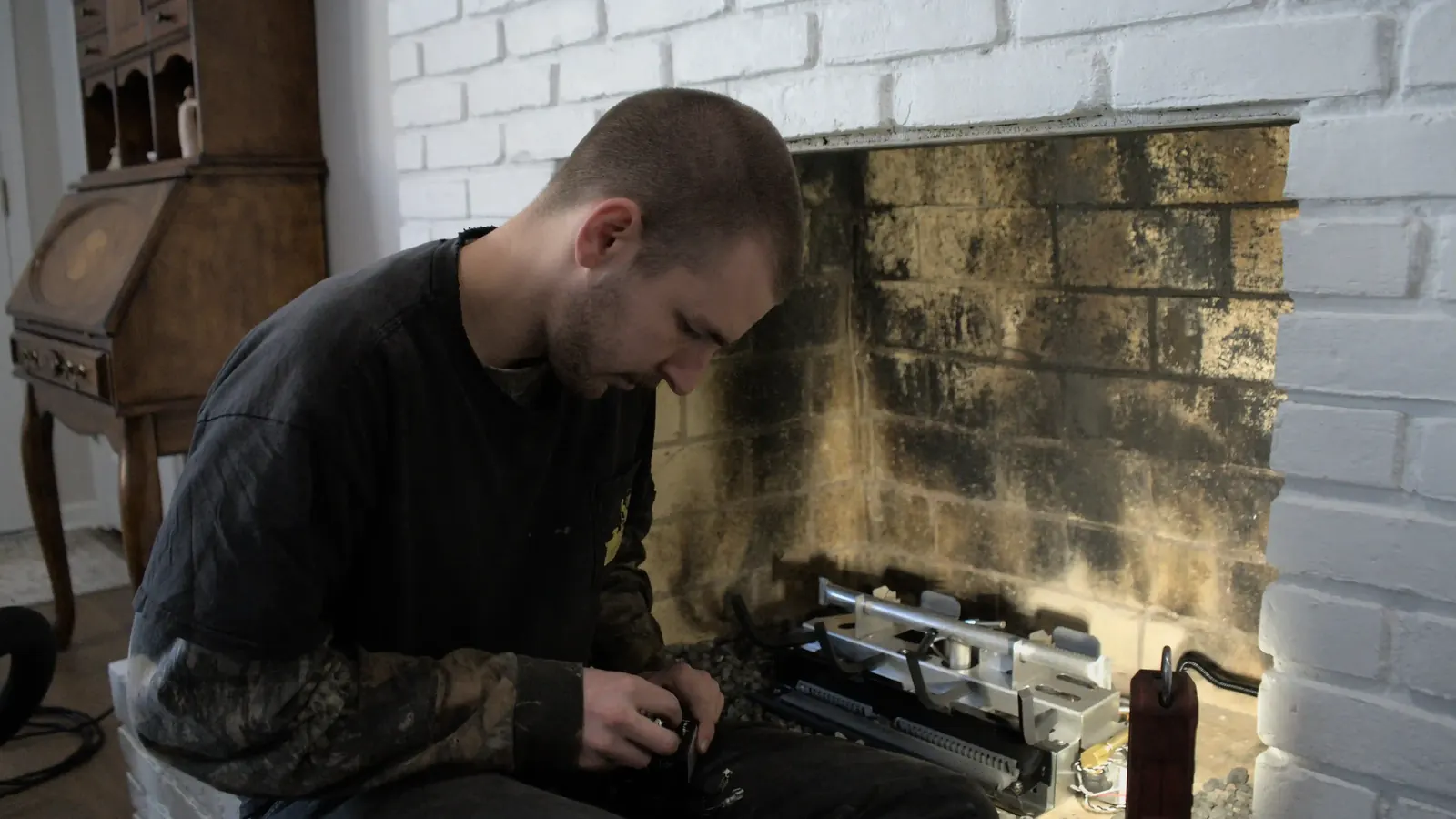 A technician servicing a gas burner assembly at a brick fireplace
